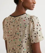 Blusa de crepé color beige con estampado de lunares de colores, cuello redondo con ribete negro, manga corta abullonada, bolsillo de parche en el pecho y bajo mullet con aberturas laterales.