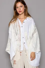 Camisa blanca de manga larga con detalles de encaje y crochet, cuello con escote en V y cierre frontal con botones. Diseño oversized con dobladillo asimétrico.