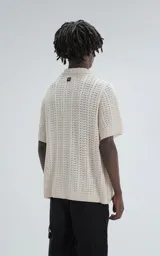 Camisa de tejido de punto color beige, manga corta, con cuello polo y cierre frontal de botones. Presenta un calce regular y un patrón calado en zigzag.