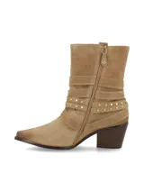 Bota corta de cuero gamuza color beige, con taco de madera y punta fina. Presenta tres tiras con hebillas metálicas plateadas y tachas pequeñas alrededor del tobillo.