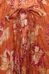 Camisa arrugada de ramio color naranja con estampado floral en tonos rosados y blancos. Tiene cuello redondo con escote pico ajustable con lazadas, mangas 3/4 y bajo asimétrico con aberturas laterales.