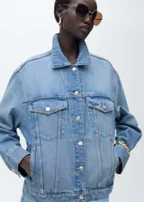 Chaqueta de jean celeste de corte oversize, con cuello camisero, cierre frontal con botones metálicos, dos bolsillos de parche con botón en el pecho y dos bolsillos laterales.