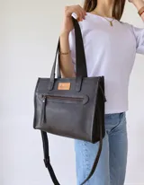 Cartera tipo tote bag de cuero color negro, con doble asa corta de mano y correa larga regulable y extraíble para usar al hombro o cruzada. Tiene un bolsillo frontal con cierre y un bolsillo interno.
