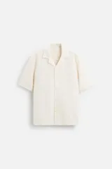 Camisa color crudo de corte regular fit, confeccionada en tejido con textura. Tiene cuello bowling, manga corta, bajo con aberturas laterales y cierre frontal con botones.