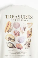 Remera blanca de algodón con cuello redondo y estampado pequeño en el pecho con la frase "Treasures of the Tides" y un dibujo de una concha. Corte holgado.