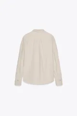 Camisa de manga larga color crudo, confeccionada en hilatura de algodón. Tiene cuello solapa, bolsillo de plastrón en el pecho, bajo redondeado y cierre frontal con botones.
