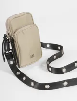 Porta celular tipo bandolera de color beige, con dos compartimentos frontales con cierre. Cuenta con una correa ajustable negra adornada con ojalillos metálicos plateados.