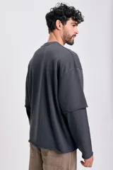 Sudadera de manga larga color gris oscuro con efecto desgastado y dobladillo sin rematar en el bajo. Presenta un diseño de doble manga, donde la manga interior es visible en el puño. Estampado frontal en color rosado con texto temático.