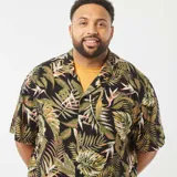 Camisa de manga corta con estampado de hojas tropicales en tonos verdes y marrones sobre fondo negro. Tiene cuello clásico, corte recto y cierre frontal con botones.