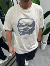 Remera beige de algodón con cuello redondo y estampado frontal de un rostro en tonos grises y texto "FUTUREPROOF".