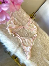 Conjunto de lencería de encaje floral en tonos rosa y crema, compuesto por un corpiño triangular sin aro con breteles finos regulables y una tanga regulable.