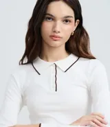 Blusa blanca de punto con cuello polo y mangas largas. Presenta detalles contrastantes en negro en el cuello, la abertura frontal y el dobladillo.