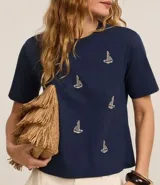Blusa azul marino de algodón con cuello redondo y mangas cortas. Presenta bordados de barcos de vela en la parte delantera.