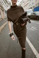 Vestido negro de tejido de punto, corte midi y silueta holgada, con cuello alto acanalado y mangas cortas tipo murciélago. Se ajusta a la cintura con un cinturón fino de efecto piel negro con hebilla dorada en forma de C.