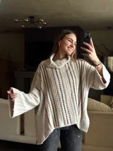 Sweater tejido color beige con cuello alto y mangas largas. Presenta aberturas verticales con detalles en marrón y negro.