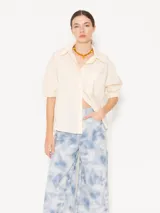 Camisa color crema de poplin liso, con acceso frontal a través de cartera oculta con seis botones forrados en tela base y un botón petra en el pie de cuello. Las mangas cortas finalizan en puño con elástico. Incluye un bolsillo plaqué con atraques triangulares para refuerzo y bordado marcario JCH en el lateral inferior.