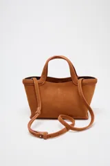 Bolso mini shopper color marrón de serraje, con asa de mano y asa bandolera extraíble. Tiene detalle de pespunte en el corte, bolsillo interior y cierre mediante mosquetón.