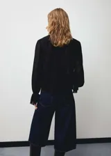 Blusa negra de tejido fluido con cuello alto, cierre frontal oculto con botones, mangas largas con puños elásticos y volantes.