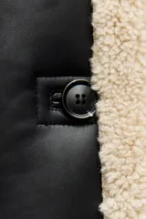 Cazadora de cuero sintético negro con interior de borreguillo color crema. Tiene cuello solapa, mangas largas, bolsillos delanteros y cierre con botones.