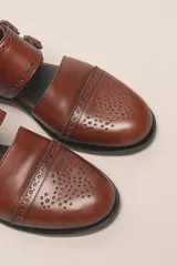 Zapato bajo de cuero negro, estilo monk strap con dos hebillas ajustables sobre el empeine. Presenta detalles de brogue (perforaciones) en la puntera y alrededor del borde superior. Tiene un taco bajo y ancho.
