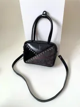 Cartera rígida negra de cuero con tachas plateadas y tres combinaciones de cuero diferentes que aportan textura. Tiene herrajes y cierre metálico plateado marca YKK. Cuenta con un asa fina para llevar al hombro o en la mano y una correa larga extra regulable.