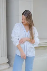 Camisa blanca con bordado de flores, cuello en V, cierre frontal con botones, mangas largas con puños elásticos y detalles de encaje.