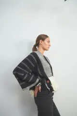 Chaqueta de punto en color negro con detalles en blanco, de frente abierto y diseño holgado. Presenta paneles frontales con patrón de zigzag y terminación de flecos en el ruedo, mientras que las mangas exhiben un motivo de líneas punteadas horizontales.