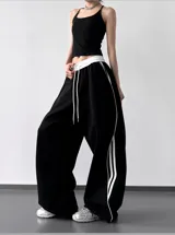 Pantalón deportivo negro de calce extra amplio (wide leg) con cintura elástica blanca y cordón ajustable blanco. Presenta tres tiras verticales blancas a lo largo de las costuras laterales.