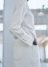 Camisa blanca de poliéster con cuello camisero, cierre frontal con botones y mangas largas con puños abotonados. Diseño recto y tejido fluido.