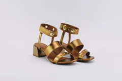 Sandalias plateadas con dos tiras horizontales sobre el pie y una tira vertical que se une a una pulsera con hebilla en el tobillo. La pulsera tiene ojales y tachas combinadas. El taco es cuadrado y de 5 cm.