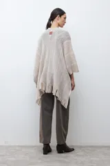 Ruana de tejido de punto en tonos beige y blanco, con patrón de espiga. Presenta cuello en V, mangas amplias, dos bolsillos frontales y flecos en el dobladillo.