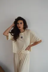 Remera de manga corta color crema, con textura acanalada y cuello redondo. Presenta un pequeño parche bordado con un diseño floral o geométrico en el pecho.