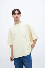 Remera color crema de algodón con calce boxy, manga corta y cuello redondo. Presenta un pequeño estampado con texto en el pecho y un diseño de texto más grande en la espalda.