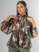 Camisa de seda italiana con estampado floral en tonos oscuros (verde oliva, rosado, beige) y volados frontales. Presenta cuello alto con lazo ajustable en la espalda y mangas abullonadas con puños ajustados.