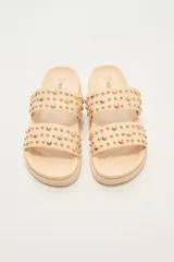 Sandalia tipo slide de color crudo, con dos tiras anchas adornadas con tachas doradas redondas de diferentes tamaños. La suela es gruesa y de goma.