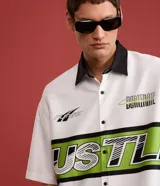 Camisa blanca oversized de tricolina con cuello negro contrastante, mangas cortas y estampado con la palabra "Hustle" en verde y negro.