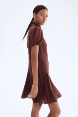 Vestido corto color marrón oscuro, con cuello alto y lazo frontal. Presenta mangas cortas abullonadas y una falda con vuelo y pliegues.