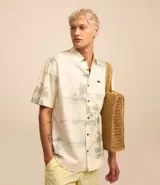 Camisa de manga corta con estampado de palmeras y motivos playeros en tonos celeste y verde sobre fondo blanco.
