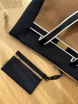 Bolso tipo tote de neopreno color negro y marrón, con cierre superior, bolsillo frontal marrón con logo rojo, bolsillo interno y externo con cierre, doble asa de mano marrón con detalles blancos y opción de correa larga.