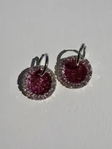 Par de aros colgantes con argolla de acero quirúrgico y dije circular de resina color fucsia con glitter y borde de strass plateado.
