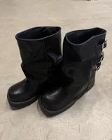 Bota corta negra de estilo motociclista, caña arrugada y suela plataforma gruesa. Presenta tres hebillas metálicas ajustables en el lateral exterior.