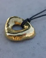 Collar con dije dorado irregular con agujero central, colgando de cordón negro ajustable.