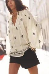 Blusa color crudo de cuello redondo con puntilla, frunces y cordón. Escote pico y manga larga abullonada. Detalle de bordados a contraste en negro.