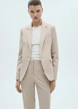 Blazer color beige con solapas de muesca, cierre frontal con un botón, hombros estructurados, corte entallado, forro interior y dos bolsillos de ribete en la parte delantera.