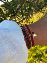 Pulsera tejida con hilo rojo y dijes colgantes: medallita dorada con mensaje Good Luck, perlita de río, perla de acero dorada y cuenta de jade roja.