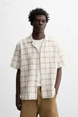 Camisa de manga corta color crudo con estampado de cuadros en tonos grises y marrones. Tiene cuello bowling, corte relaxed fit, aberturas laterales en el bajo y cierre frontal con botones.