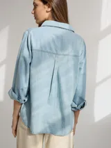 Camisa celeste de denim suave, corte relajado, con cuello clásico, cierre frontal con botones marrones, manga larga con puños abotonados, dos bolsillos de parche en el pecho y ruedo redondeado.