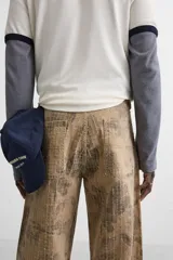 Pantalón de jean color beige con estampado abstracto floral en tonos marrones, de corte amplio, con bolsillos delanteros y traseros, y detalle de tira multifunción en la pierna. Cierre frontal con cremallera y botón.