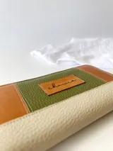 Billetera grande con cierre metálico, confeccionada en cuero con diseño de bloques de color: verde oliva texturizado, marrón claro liso y blanco/crema texturizado. Posee un monedero interno y un tirador de cuero marrón claro.
