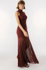 Vestido largo color beige, con cuello alto y sin mangas. La parte superior es ajustada al cuerpo y la falda es amplia y fluida, con paneles traslúcidos.